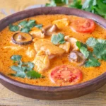 Close de uma tigela de curry vermelho decorada com folhas de coentro verde e rodelas de pimenta vermelha.