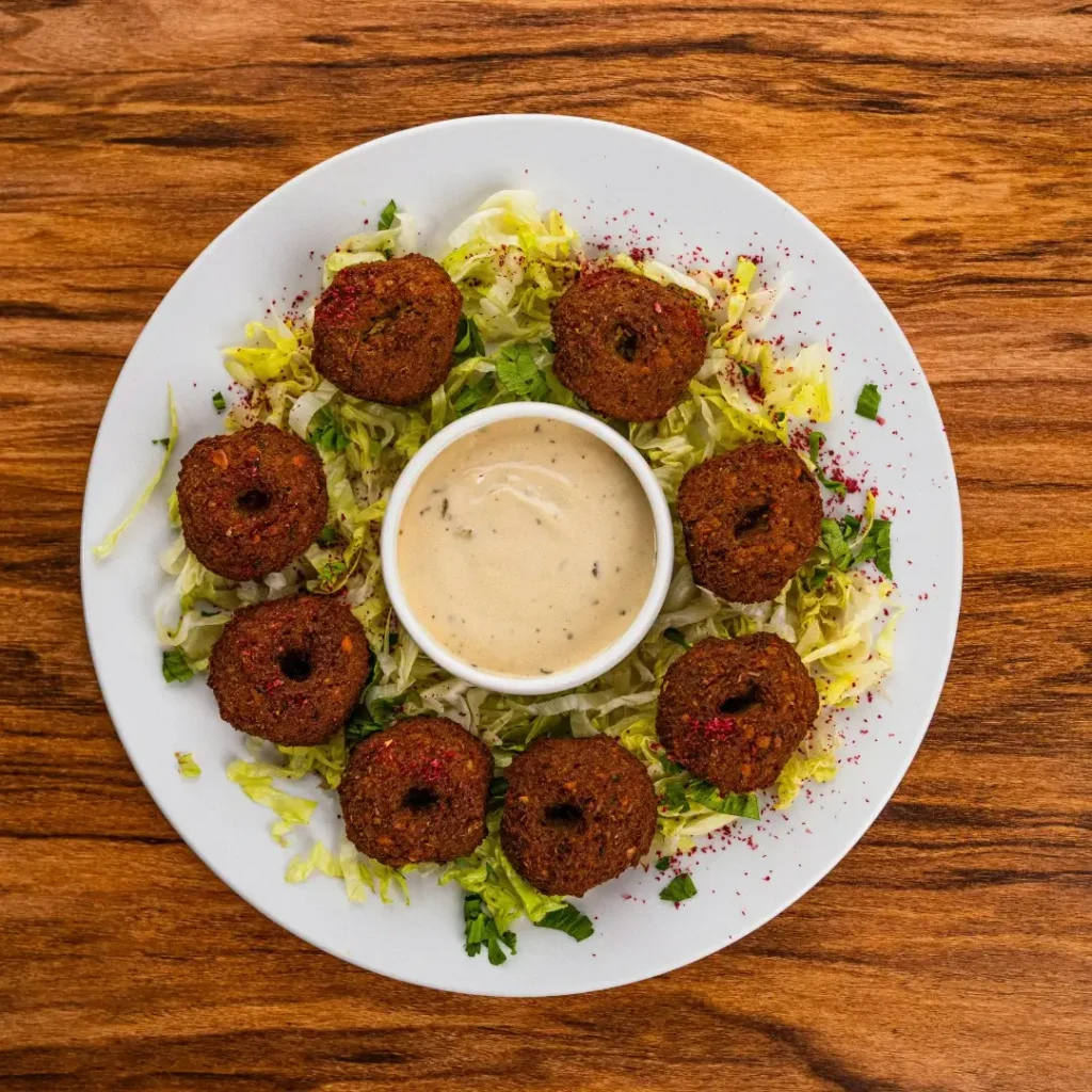 Prato com vários bolinhos de falafel dourados, servidos com salada de tomate, pepino e um pote de molho branco.