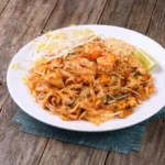 Prato de Pad Thai com camarões grandes por cima, amendoim triturado ao lado e fatias de limão