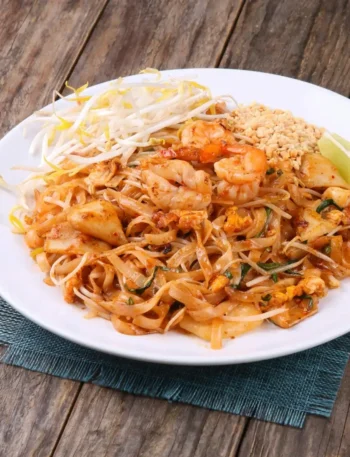 Prato de Pad Thai com camarões grandes por cima, amendoim triturado ao lado e fatias de limão