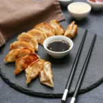 Prato de gyoza cozido no vapor e frito, servido com um pequeno pote de molho escuro e palitinhos de madeira.