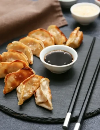 Prato de gyoza cozido no vapor e frito, servido com um pequeno pote de molho escuro e palitinhos de madeira.