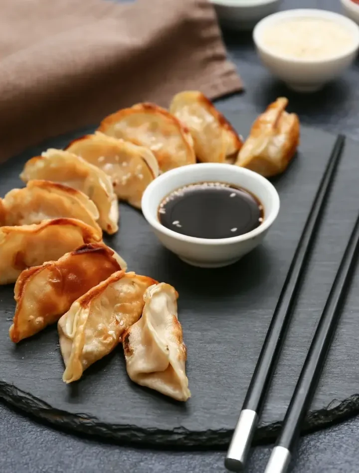 Prato de gyoza cozido no vapor e frito, servido com um pequeno pote de molho escuro e palitinhos de madeira.