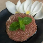 Prato de kibe cru decorado artisticamente com folhas de hortelã verde e muito azeite de oliva por cima.