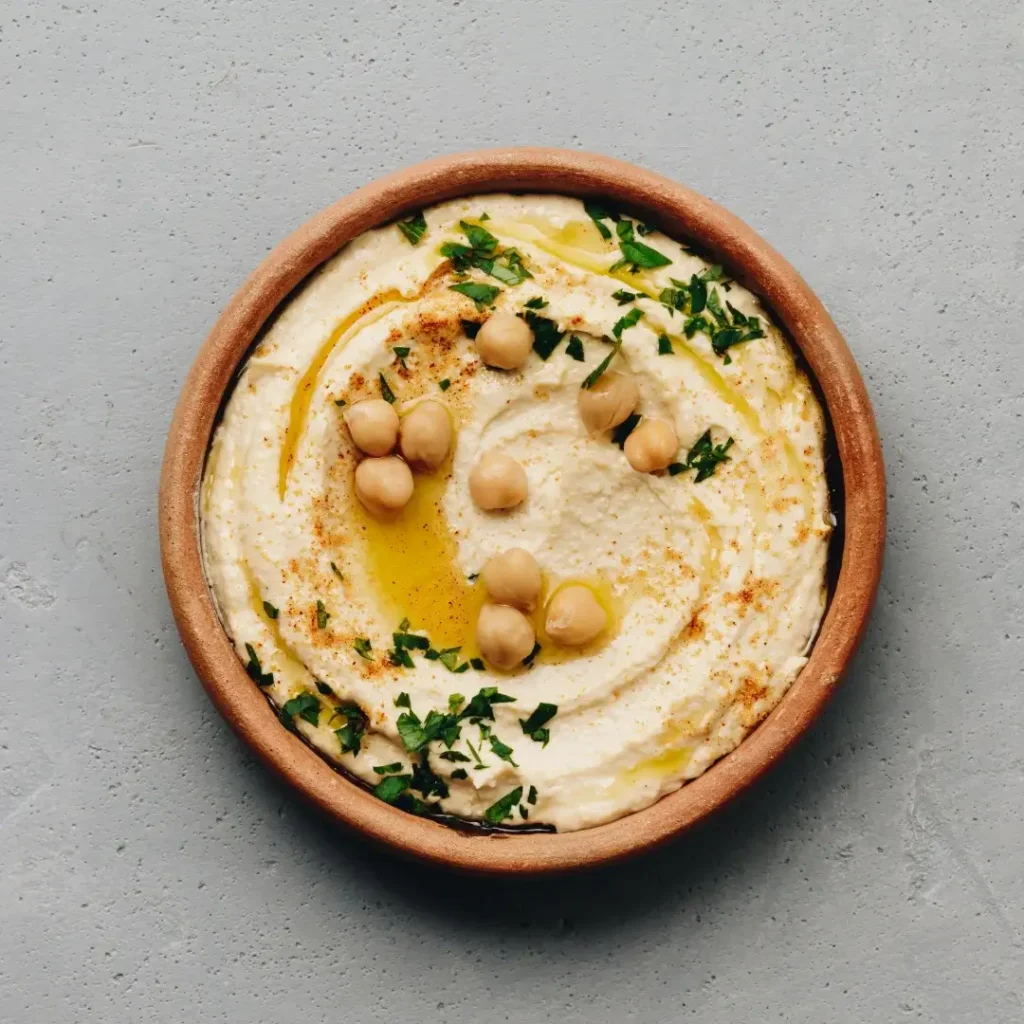 Uma tigela de cerâmica com homus bege liso, decorada com azeite, páprica e grãos de bico por cima.
